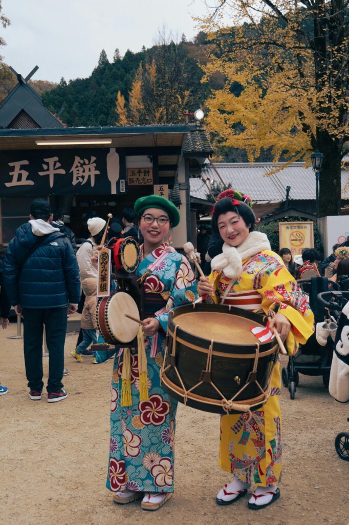 Two women in kimonos playing traditional drums at a Japanese festival outdoors.